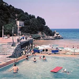 A stretch of coast of the island of Elba. In the foreground a swimming pool with bathers. On the left a staircase leads to a terrace